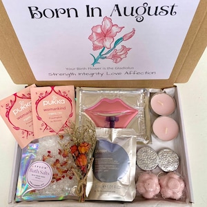 May include: A gift box with the text "Born In August" and a floral illustration. Inside are pink tea packets, bath salts, candles, and other self-care items. The box includes a lip-shaped item and small, round, silver-colored items.