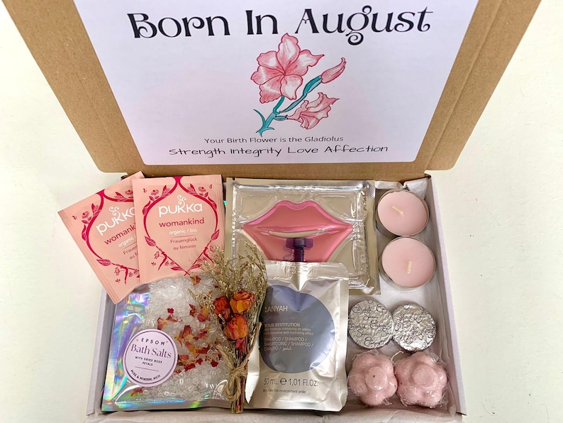 May include: A gift box with the text "Born In August" and a floral illustration. Inside are pink tea packets, bath salts, candles, and other self-care items. The box includes a lip-shaped item and small, round, silver-colored items.