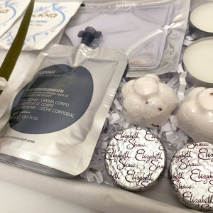 May include: A gift box filled with spa products. Includes white tea light candles, wrapped soaps, and foil-wrapped chocolates. A packet of body cream is also visible, along with a sprig of white flowers.