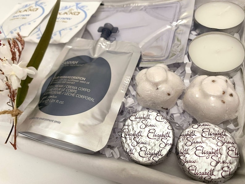 May include: A gift box filled with spa products. Includes white tea light candles, wrapped soaps, and foil-wrapped chocolates. A packet of body cream is also visible, along with a sprig of white flowers.