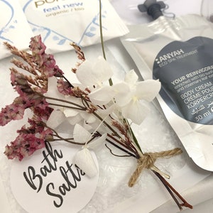 May include: A spa-themed product arrangement featuring a small bouquet of dried flowers and a tag that reads "Bath Salts." A packet of body cream with text is visible, along with other spa products. The bouquet is tied with twine.