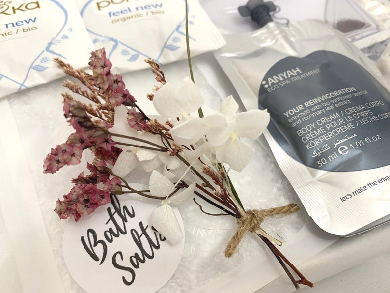 May include: A spa-themed product arrangement featuring a small bouquet of dried flowers and a tag that reads "Bath Salts." A packet of body cream with text is visible, along with other spa products. The bouquet is tied with twine.