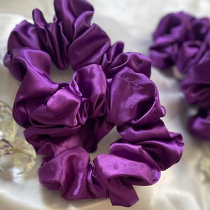 May include: Several vibrant purple satin scrunchies are arranged on a white surface. The scrunchies are bunched and gathered, showcasing the fabric's sheen. Clear, decorative crystals are scattered around the scrunchies, adding a touch of sparkle.