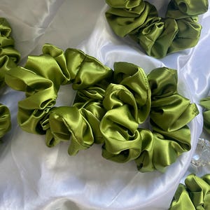 Peut inclure: Plusieurs chouchous en satin vert olive sont disposés sur un tissu blanc et soyeux. Les chouchous sont froncés et froissés, révélant une texture lisse et brillante. L'image met en valeur la couleur verte vibrante et l'éclat du tissu. Accessoires pour cheveux.