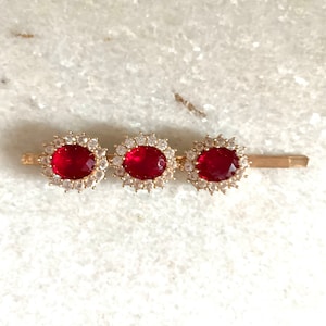 May include: Gold hair clip with three red oval-shaped gemstones surrounded by clear rhinestones.