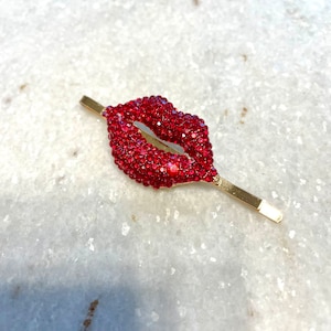May include: A gold-toned hair clip featuring a sparkling red lips design. The lips are encrusted with small, vibrant red rhinestones. The clip is a stylish accessory for hair styling.