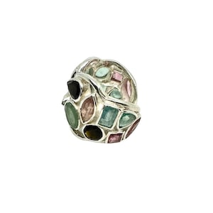 Op de afbeelding: Een zilveren ring met een cluster van kleurrijke edelstenen. De ring is bezet met verschillende vormen en maten van groene, roze en zwarte stenen.