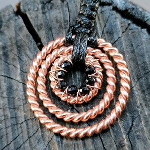 Peut inclure: Un collier en fil de cuivre avec un pendentif circulaire composé de trois cercles concentriques de fil torsadé. Le cercle central est rempli de pierres précieuses noires.