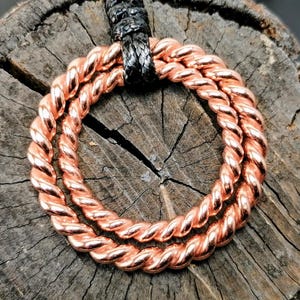Op de afbeelding: Een koperen hanger met een gevlochten ontwerp, bestaande uit drie in elkaar verweven cirkels. De hanger is bevestigd aan een zwart koord.