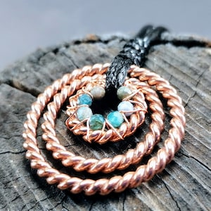 Puede incluir: Un collar con colgante de color cobre con un diseño en espiral. El colgante presenta tres anillos concéntricos retorcidos, con un anillo interior más pequeño que contiene cuentas azules y verdes. El collar tiene un cordón negro.