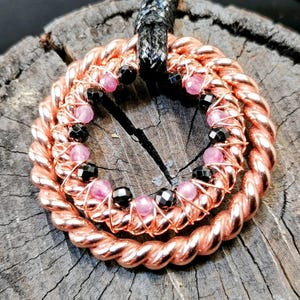 Puede incluir: Un collar de alambre de cobre con cuentas rosas y negras. El collar tiene forma de círculo y está hecho de dos capas de alambre. Las cuentas están espaciadas uniformemente alrededor del círculo.