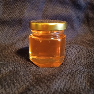 May include: A small glass jar with a gold lid filled with honey. The jar is on a brown fabric background.
