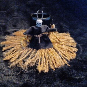 May include: A Halloween figurine of a skull-headed figure in a black robe, standing on a skirt of tan twine. A small black cauldron is behind the figure. The background is a dark, textured fabric.