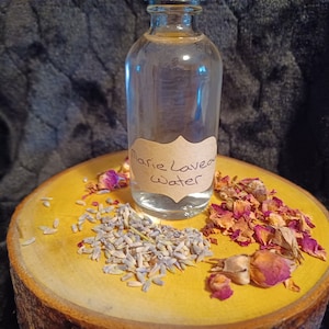 May include: A clear glass bottle with a brown label that reads "Marie Laveau Water" on a wooden surface with dried lavender and rose petals.