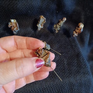 May include: A close-up of a hand holding a small, dried, brown, and black dog chew with a thin, gold wire attached. The chew is on a black fabric background.