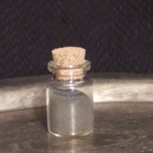 Puede incluir: Una pequeña botella de vidrio con un tapón de corcho, llena de un polvo gris oscuro.