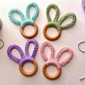 May include: Four wooden rings with macrame bunny ears in light blue, sage green, purple, and soft pink. Each ring features a smooth, circular wooden base with knotted cord forming two upright loops that resemble rabbit ears in pastel colors.