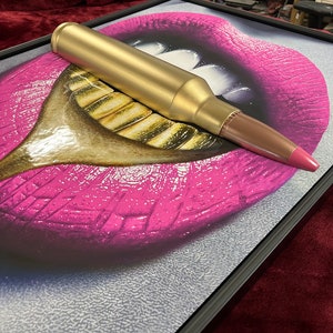 May include: A framed artwork featuring a pop art style image of pink lips with gold teeth. A gold-colored bullet is placed on top of the artwork, with a pink tip. The artwork is in a black frame.
