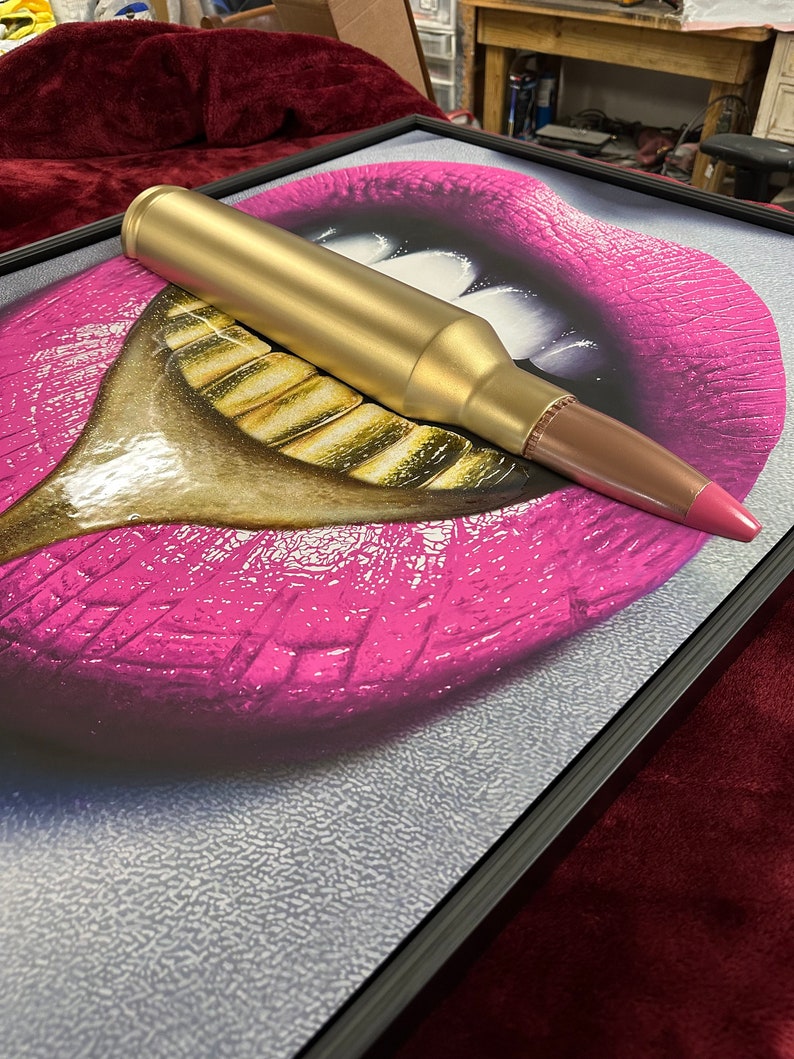 May include: A framed artwork featuring a pop art style image of pink lips with gold teeth. A gold-colored bullet is placed on top of the artwork, with a pink tip. The artwork is in a black frame.