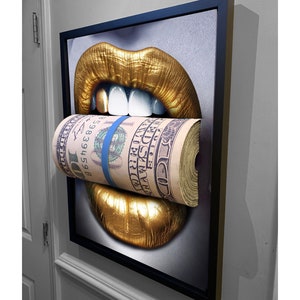 May include: Framed artwork depicting golden lips with a roll of US hundred-dollar bills held between them, secured with a blue band. The artwork is in a black frame and mounted on a wall.
