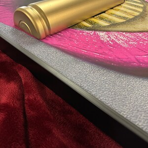 May include: A gold-colored, bullet-shaped object rests on a framed artwork featuring a close-up of a mouth with bright pink lipstick. The artwork is framed with a silver textured border, and a burgundy plush fabric is in the foreground.