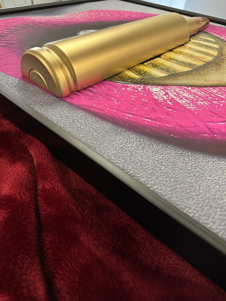 May include: A gold-colored, bullet-shaped object rests on a framed artwork featuring a close-up of a mouth with bright pink lipstick. The artwork is framed with a silver textured border, and a burgundy plush fabric is in the foreground.