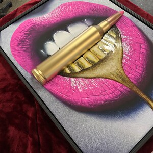 May include: Framed artwork featuring a close-up of pink lips with white teeth and a gold-colored spoon. A gold bullet rests across the lips and spoon. The artwork is framed in a black frame.