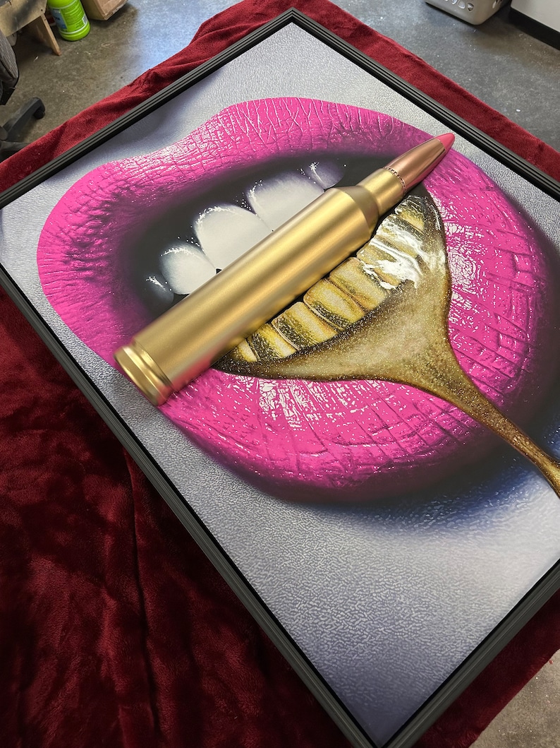 May include: Framed artwork featuring a close-up of pink lips with white teeth and a gold-colored spoon. A gold bullet rests across the lips and spoon. The artwork is framed in a black frame.