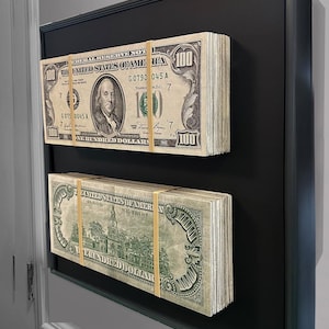 May include: Two framed stacks of US $100 bills, secured with rubber bands, are displayed against a black background. The bills feature portraits and the text "The United States of America". The frame has a dark green border.