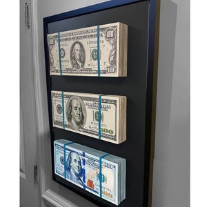 May include: Three stacks of US $100 bills are displayed in a black frame. The bills are secured with blue rubber bands. The top stack has the text "FEDERAL RESERVE NOTE" and "ONE HUNDRED DOLLARS". The middle stack has the text "UNITED STATES OF AMERICA" and "ONE HUNDRED DOLLARS". The bottom stack has the text "UNITED STATES OF AMERICA" and "ONE HUNDRED DOLLARS".