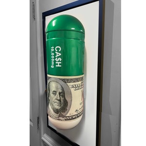 May include: A green pill-shaped sculpture with the words "CASH" and "10,000mg" printed on it. The pill is wrapped in a £100 note featuring a portrait of a man.