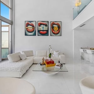 May include: A white sectional sofa with a white throw blanket in a modern living room with three red lips paintings on the wall. The paintings feature a dollar bill, a gold coin, and a cigarette.