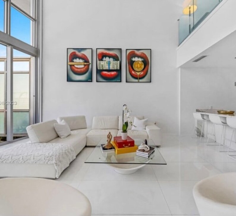 May include: A white sectional sofa with a white throw blanket in a modern living room with three red lips paintings on the wall. The paintings feature a dollar bill, a gold coin, and a cigarette.