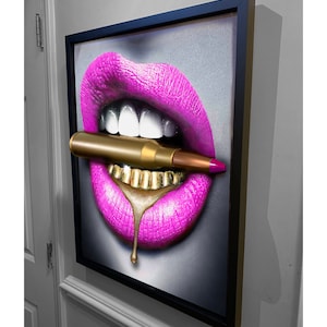May include: Framed artwork featuring a close-up of a mouth with bright pink lipstick and gold teeth. A gold bullet is held between the lips. A drop of gold liquid drips from the teeth. The artwork is framed in a black frame.