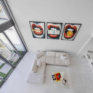 May include: A white sectional sofa with a white throw blanket and a glass coffee table with decorative items on top. Three framed prints of red lips with different objects in the mouth are hanging on the wall.