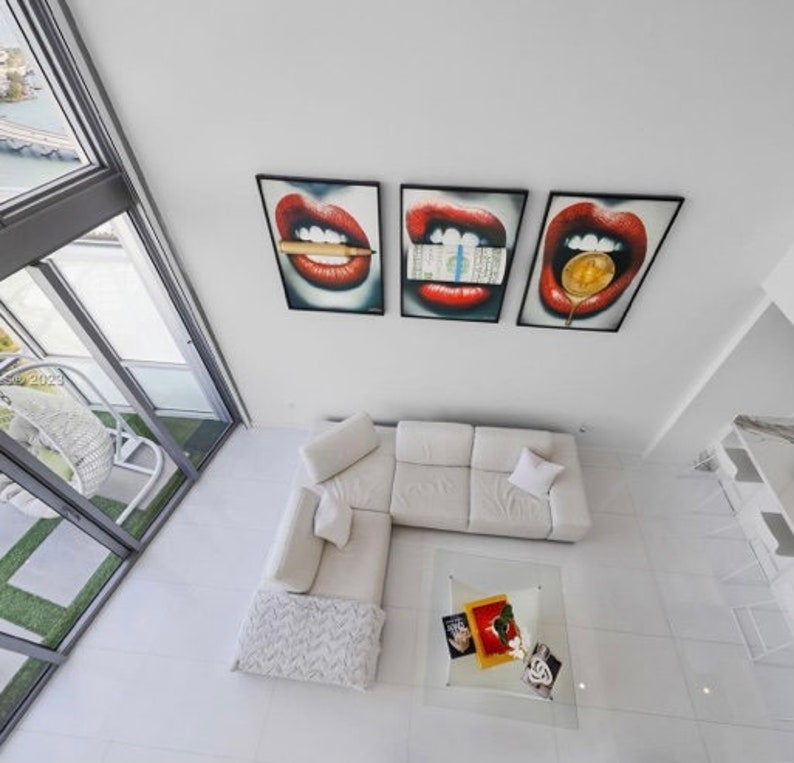 May include: A white sectional sofa with a white throw blanket and a glass coffee table with decorative items on top. Three framed prints of red lips with different objects in the mouth are hanging on the wall.