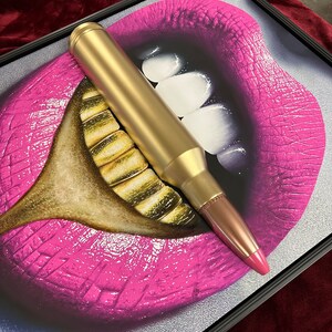 May include: Framed artwork featuring a pop art style image of a mouth with bright pink lips and gold teeth. A large gold bullet is positioned across the mouth. The artwork is framed in a black frame.