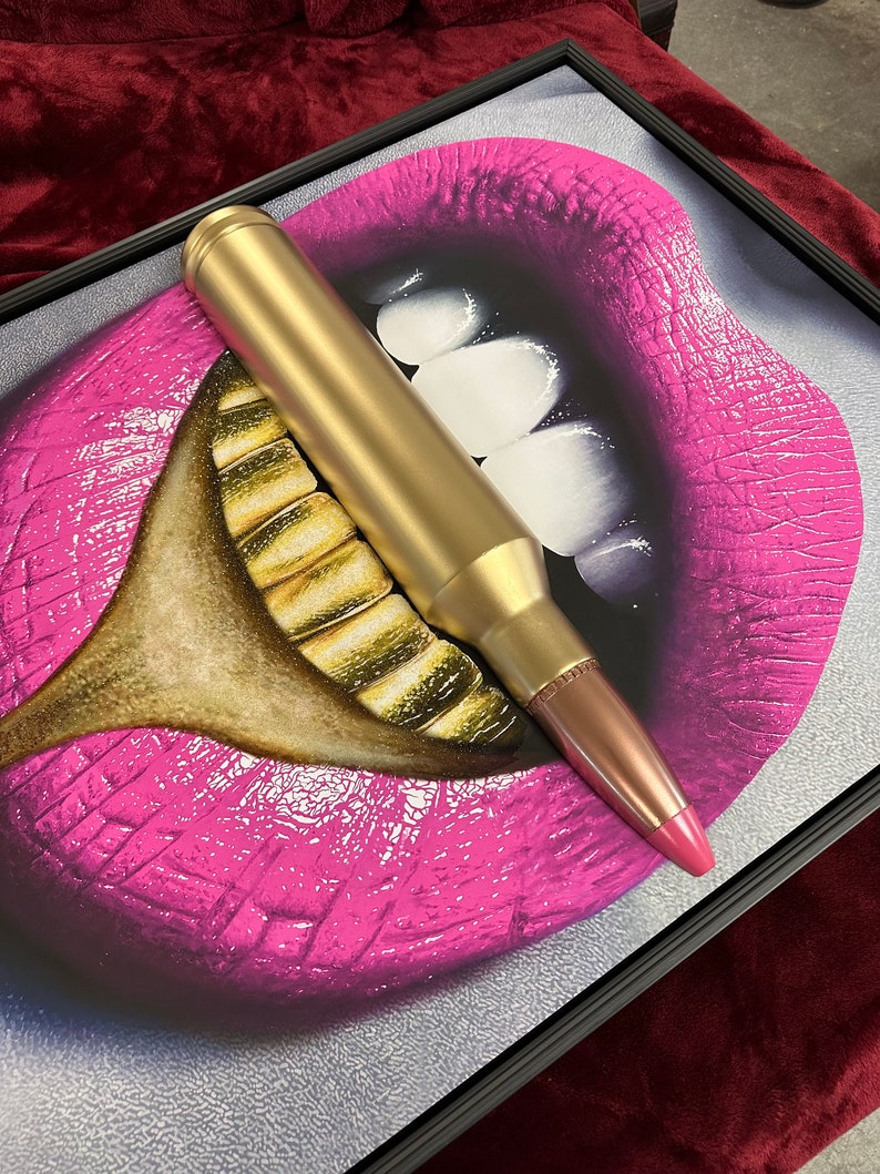 May include: Framed artwork featuring a pop art style image of a mouth with bright pink lips and gold teeth. A large gold bullet is positioned across the mouth. The artwork is framed in a black frame.
