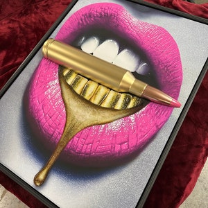May include: Framed artwork featuring a pop art style image of pink lips with gold teeth. A large, gold bullet is placed across the lips. The artwork is framed in a black frame and rests on a maroon surface.