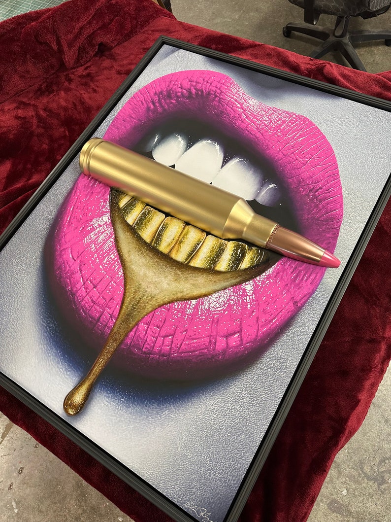 May include: Framed artwork featuring a pop art style image of pink lips with gold teeth. A large, gold bullet is placed across the lips. The artwork is framed in a black frame and rests on a maroon surface.