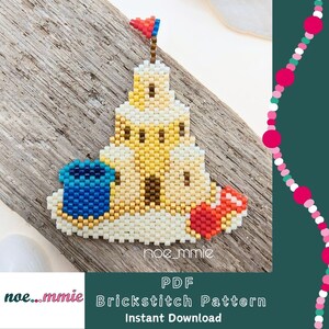 Peut inclure: Un ornement en perles en forme de château de sable avec une structure crème et jaune, un seau bleu à gauche et une pelle rouge à droite. Le château est surmonté d'un petit drapeau rouge et bleu. Le texte noe_mmie est visible en bas.