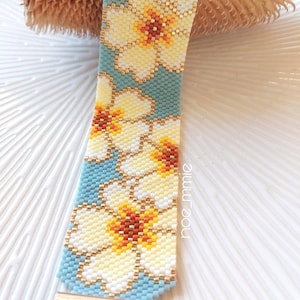 May include: A beaded bracelet with a floral pattern. The bracelet has a light blue background with white and yellow flowers with orange centres. The bracelet has a gold-coloured clasp and the word "hilos" is visible.