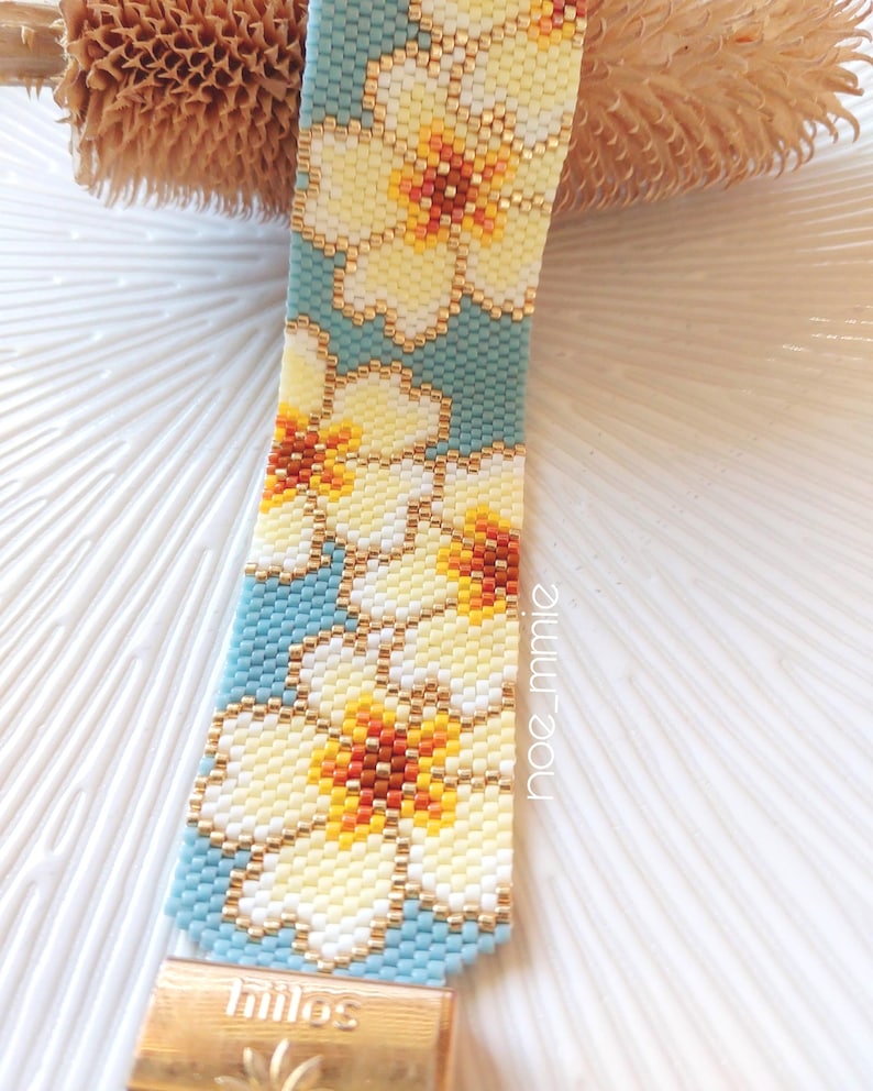 May include: A beaded bracelet with a floral pattern. The bracelet has a light blue background with white and yellow flowers with orange centres. The bracelet has a gold-coloured clasp and the word "hilos" is visible.