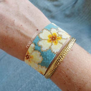 May include: A beaded cuff bracelet featuring a floral design in yellow, white, and blue, alongside a gold-tone hammered metal bracelet. The bracelets are worn on a wrist.