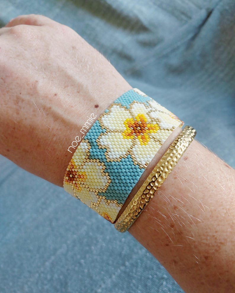 May include: A beaded cuff bracelet featuring a floral design in yellow, white, and blue, alongside a gold-tone hammered metal bracelet. The bracelets are worn on a wrist.
