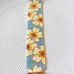 May include: A beaded bracelet with a floral design in yellow, white, and brown on a light blue backdrop. The bracelet has gold-coloured clasps and the word "hiilos" is visible. A handmade accessory.