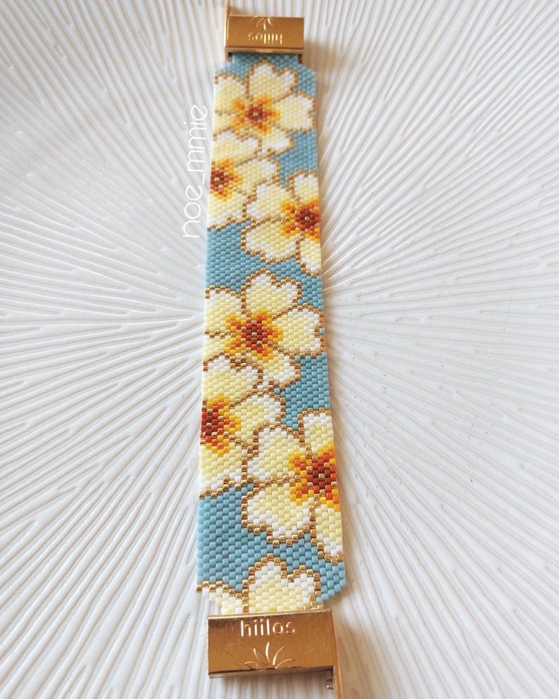May include: A beaded bracelet with a floral design in yellow, white, and brown on a light blue backdrop. The bracelet has gold-coloured clasps and the word "hiilos" is visible. A handmade accessory.