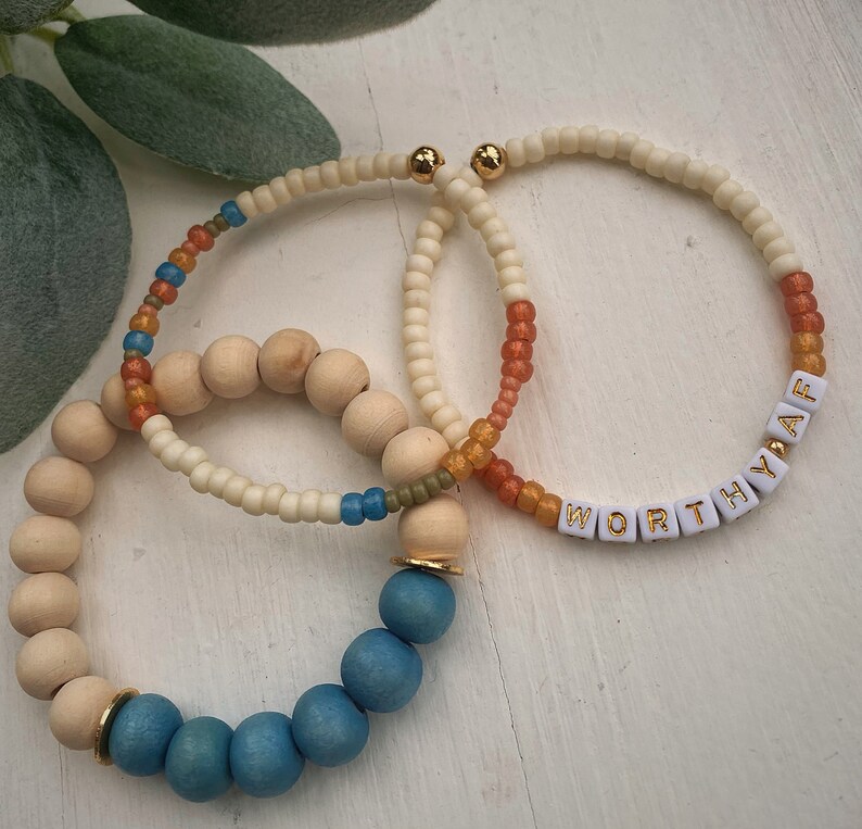Puede incluir: Tres pulseras con cuentas de madera y cuentas blancas. Una pulsera tiene cuentas azules y detalles dorados. Otra pulsera tiene cuentas naranjas y blancas. La tercera pulsera tiene cuentas blancas con la palabra "WORTHY AF" escrita en letras blancas con detalles dorados.