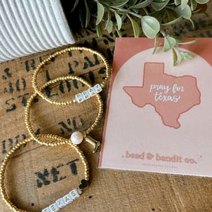 May include: Three gold beaded bracelets with white lettered beads spelling "PRAY" and "TEXAS". One bracelet has a pearl and a small charm. A card with "pray for texas" and a Texas outline is next to the bracelets.