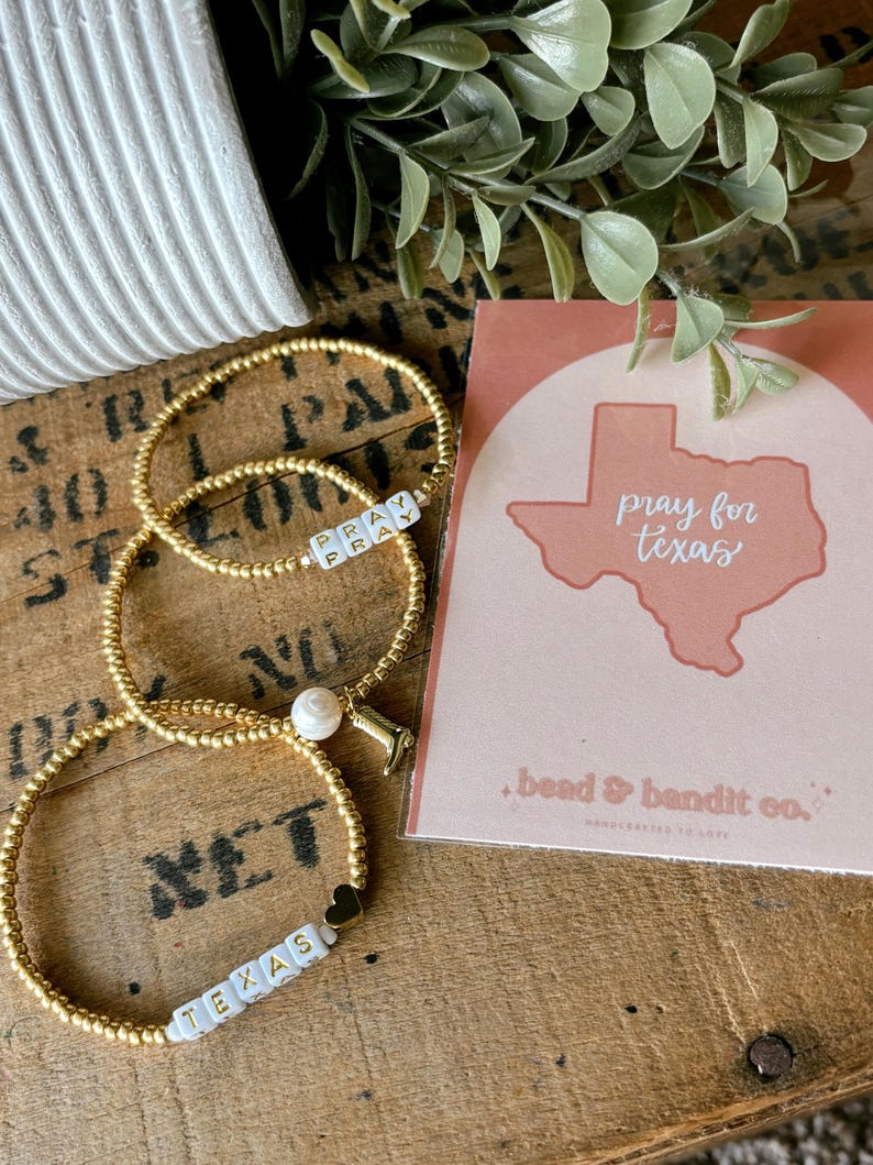May include: Three gold beaded bracelets with white lettered beads spelling "PRAY" and "TEXAS". One bracelet has a pearl and a small charm. A card with "pray for texas" and a Texas outline is next to the bracelets.